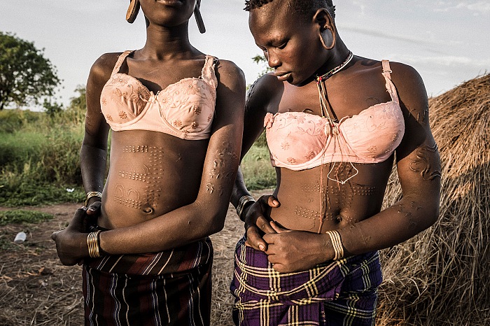 Fausto Podavini, Due ragazze di etnia Mursi si preparano a tornare al loro villaggio, dopo essere andate a prendere un po' d'acqua in un pozzo. Entrambe indossano reggiseni dati loro dai turisti occidentali, Ethiopia. Mago National Park. 2017, dalla mostra La fin d'un monde in esposizione allo Chemin des libellules nell'ambito di Festival Photo La Gacilly 2018.  Fausto Podavini.